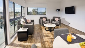 A modern living room with large windows, a flat-screen TV, and a dining area with a table and chairs.