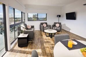 A modern living room with large windows, a flat-screen TV, and a dining area with a table and chairs.