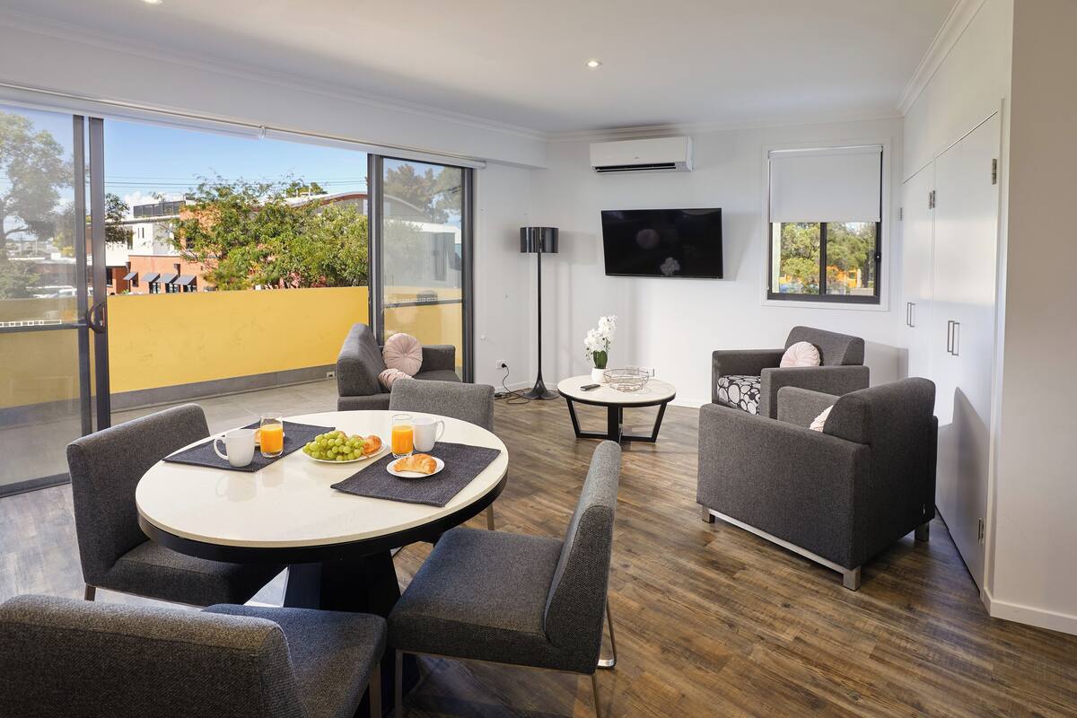 A modern living room with a round dining table, grey sofas, and a flat-screen TV mounted on the wall.