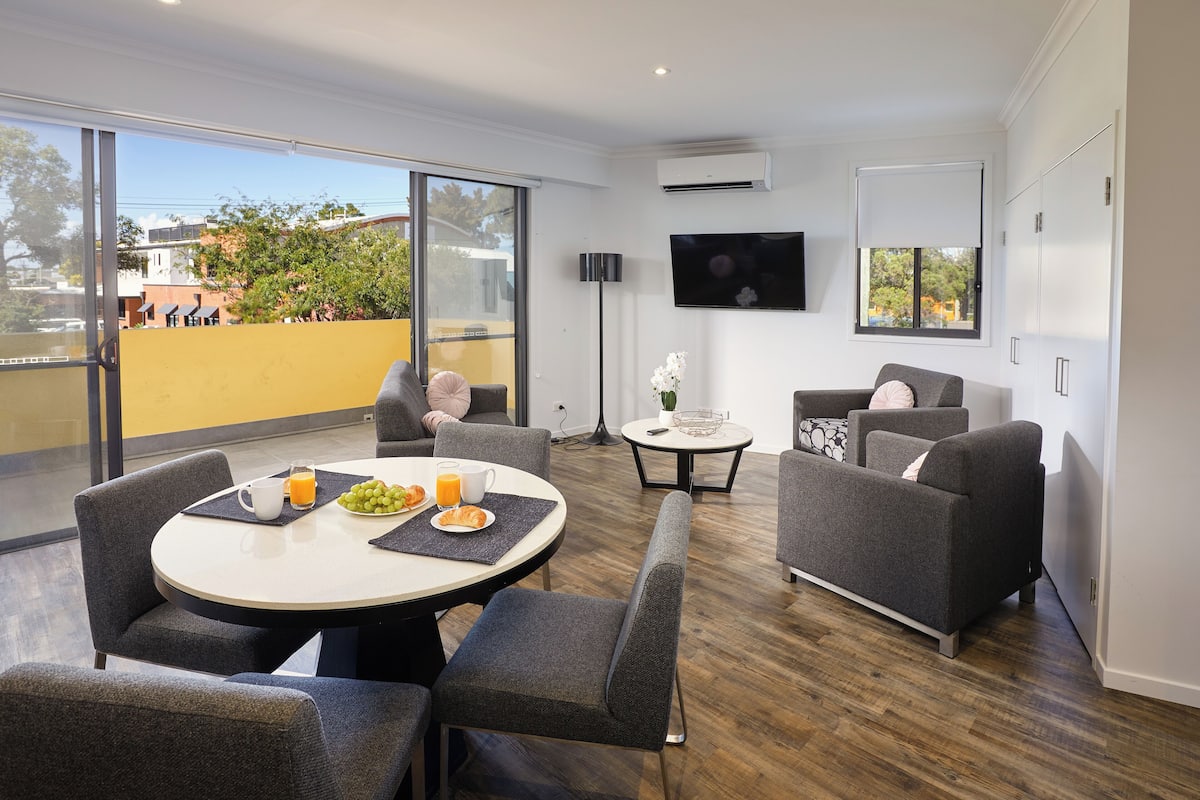 A modern living room with a round dining table, grey sofas, and a flat-screen TV mounted on the wall.