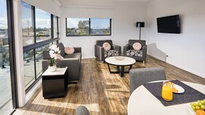 A modern living room with large windows, a flat-screen TV, and a dining area with a table and chairs. - Nouveau Serviced Apartments (Newcastle)