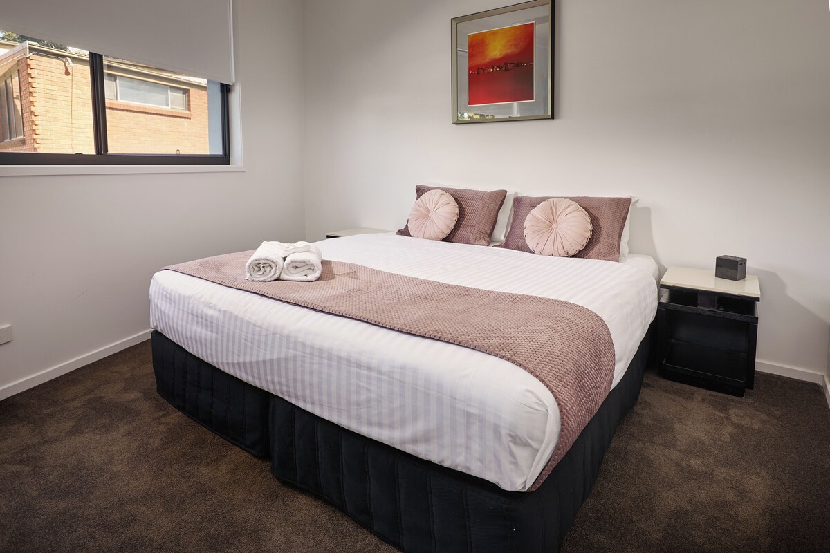 A neatly made bed with a striped bedspread and a dark skirted base, a bedside table with a lamp, and a window with a view of a brick building.