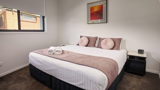 A neatly made bed with a striped bedspread and a dark skirted base, a bedside table with a lamp, and a window with a view of a brick building.