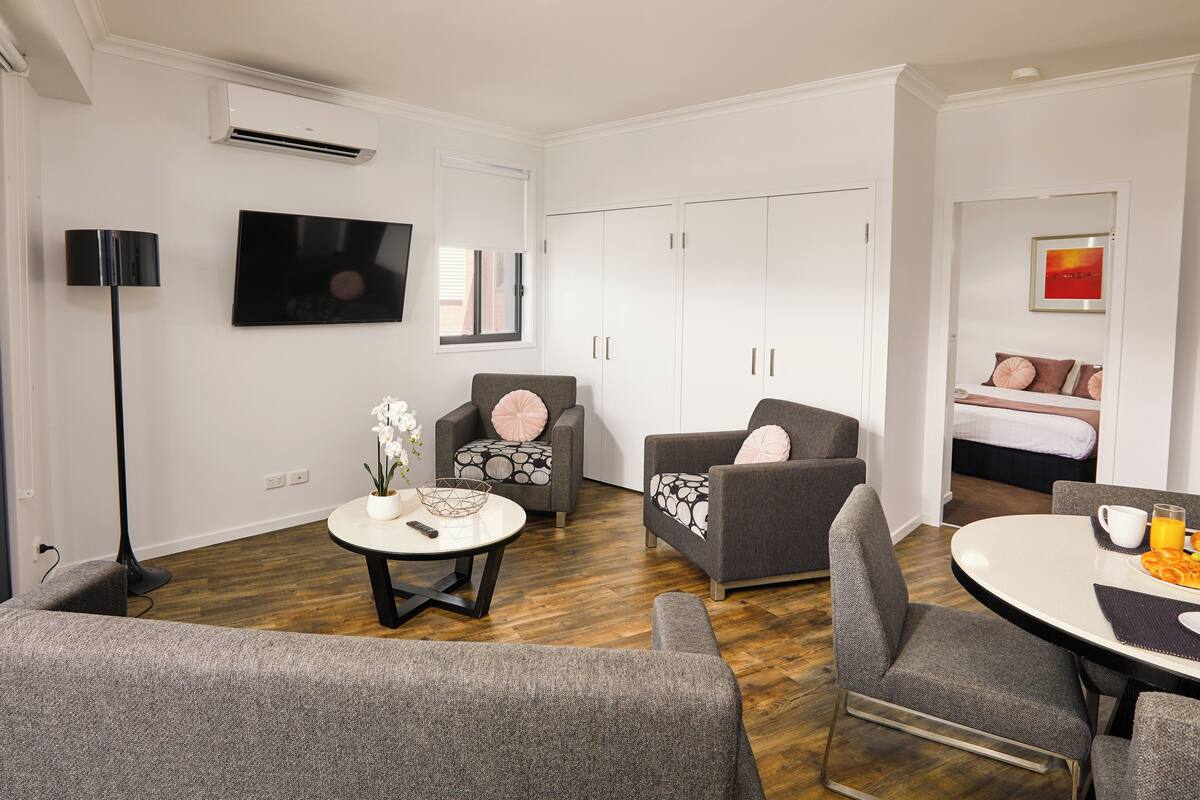 A modern living room with a flat-screen TV, grey armchairs, a round coffee table, and a dining table with breakfast items.