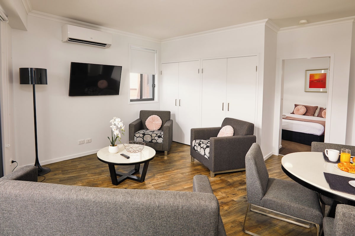 A modern living room with a flat-screen TV, grey armchairs, a round coffee table, and a dining table with breakfast items.
