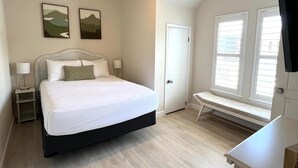 Un dormitorio con cama, ventilador de techo, escritorio y dos ventanas con persianas.