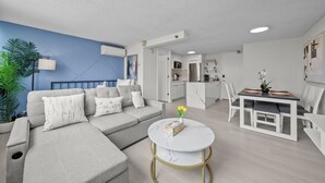 Un salon moderne avec un canapé d’angle, une table basse ronde et un coin repas comprenant une table et des chaises blanches.
