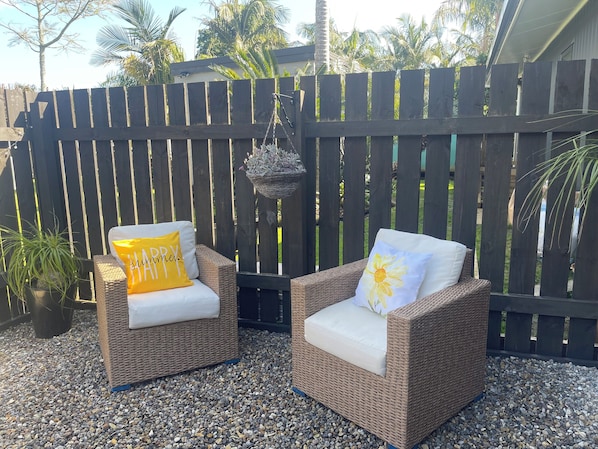 Two wicker chairs with cushions, one with a yellow pillow and the other with a white pillow with a sun design, placed on a gravel surface with a wooden fence in the background.