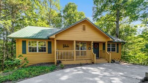 A yellow wooden cabin with a green roof, a porch, and a driveway.