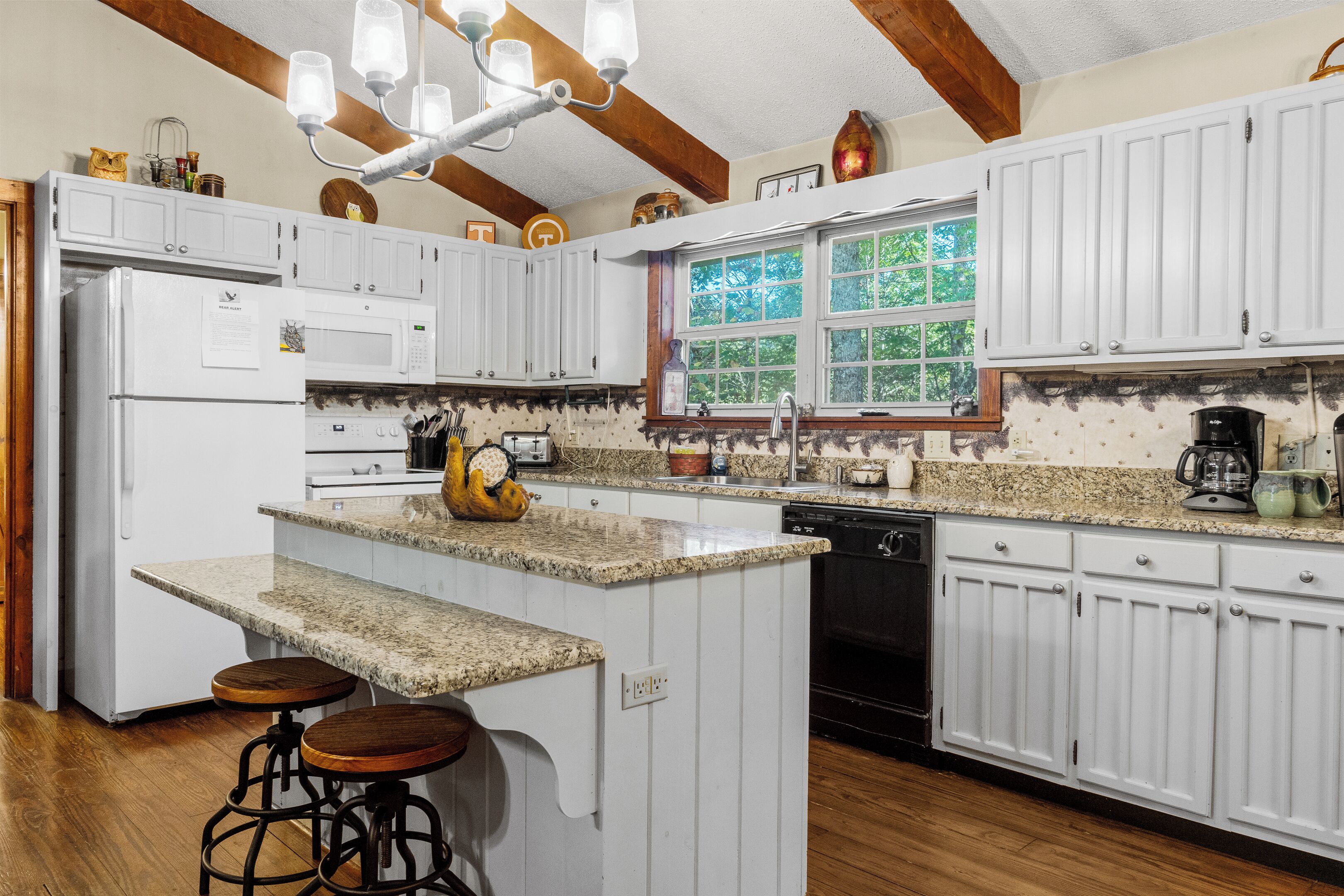 A kitchen with white cabinets, a granite countertop, a central island with stools, and a window with a view of trees.
