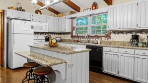 A kitchen with white cabinets, a granite countertop, a central island with stools, and a window with a view of trees.