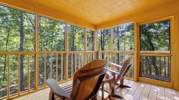 A wooden deck with large windows overlooking a forest. There are two wooden chairs and a small table.
