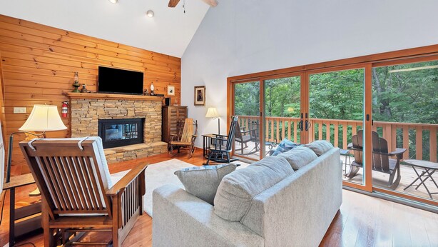 A spacious living room with a fireplace, wooden walls, a ceiling fan, and a large sliding glass door leading to an outdoor deck.