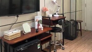 A compact room with a flat-screen TV, a desk with a chair, a small refrigerator, and a hanging clothes rack.