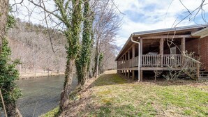 A rustic cabin with a wooden deck overlooking a river, surrounded by trees and a grassy area.