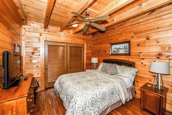 A wooden cabin bedroom with a bed, two bedside lamps, a ceiling fan, a TV, and a wooden dresser.