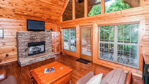 A cozy cabin interior with a stone fireplace, wooden walls, a television, and large windows overlooking a garden. - Authentic log cabin with the real feel of serenity in a Golf Course Vicinity. (Townsend)