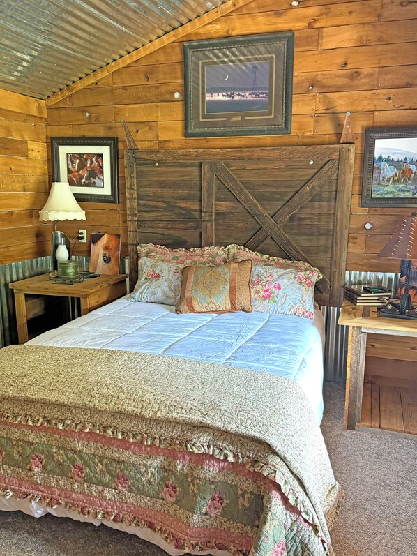 A rustic wooden room with a large bed, framed pictures on the walls, a lamp, and a small table with books and a phone. - One of a kind, property with 3 charming houses, horse facility and pets welcome. (Cle Elum)