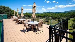 A deck with outdoor furniture and umbrellas, overlooking a green landscape with mountains.