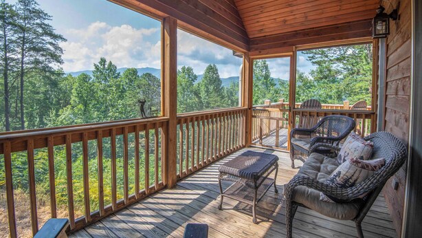 Une terrasse en bois offrant une vue sur une forêt et des montagnes. On y trouve des chaises et un banc en osier.