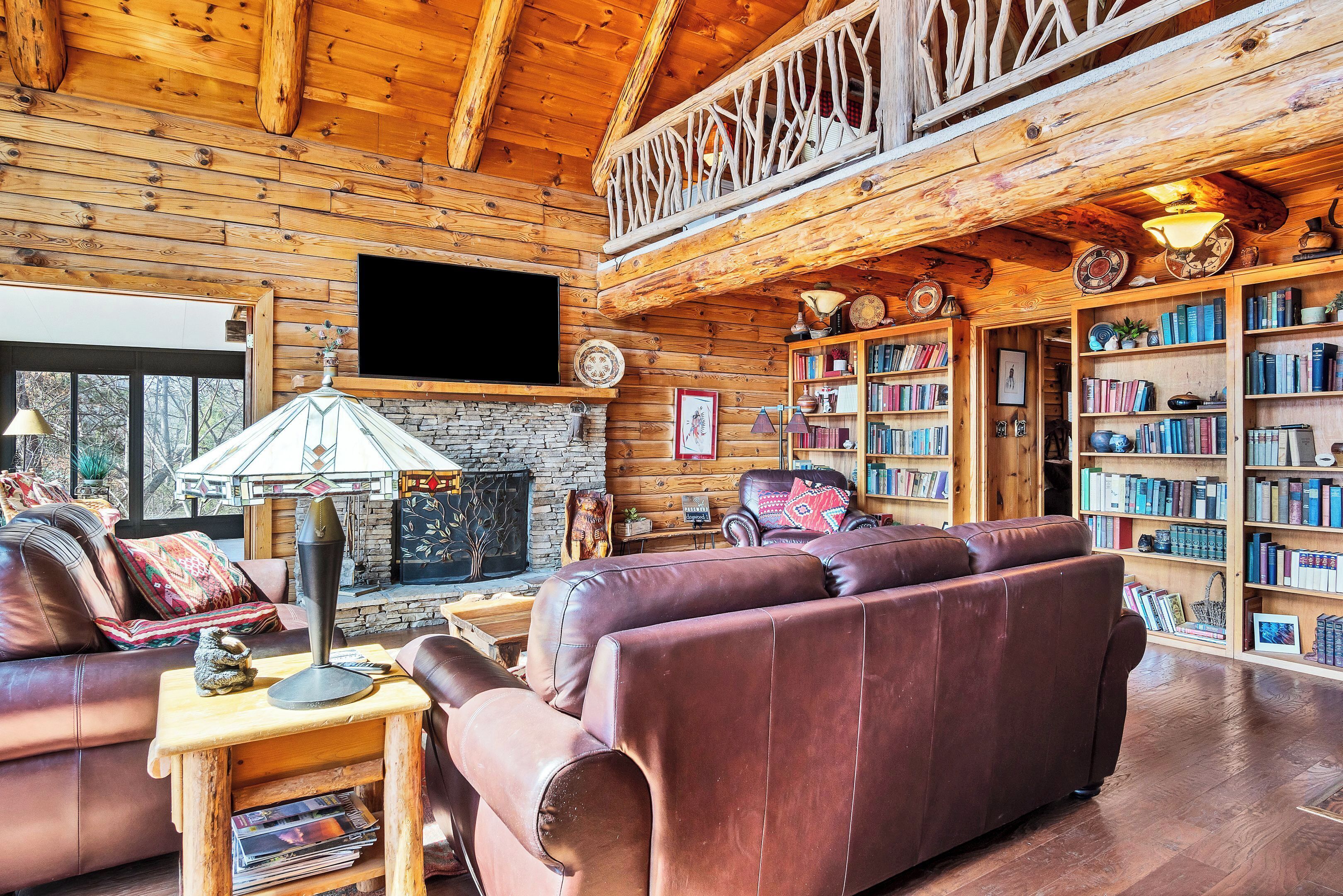 A cozy log cabin interior with a fireplace, leather sofas, a bookshelf, and a TV mounted on the wall.