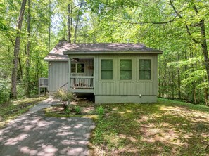 A small, single-story cabin with a porch, surrounded by dense forest.