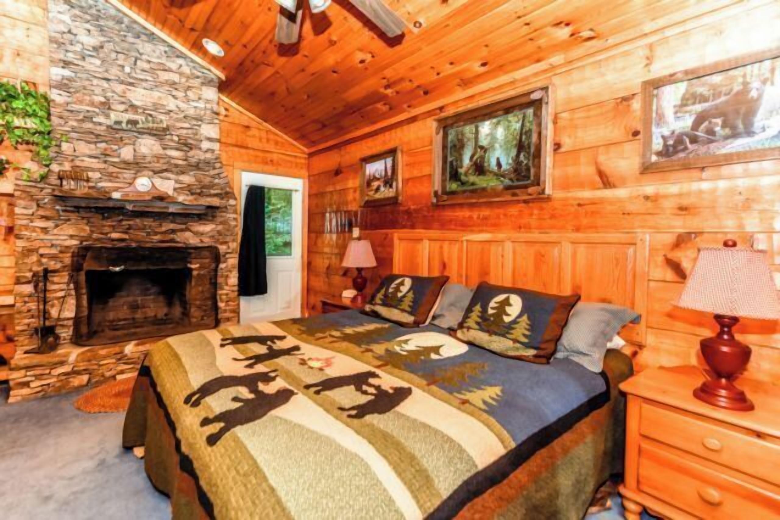 Une chambre confortable dans un chalet en rondins, avec une cheminée en pierre, un lit agrémenté d’une literie aux motifs forestiers et un tableau représentant un ours.
