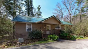 A single-story cabin with a green metal roof, a front porch, and a small front yard with a stone wall and shrubs.