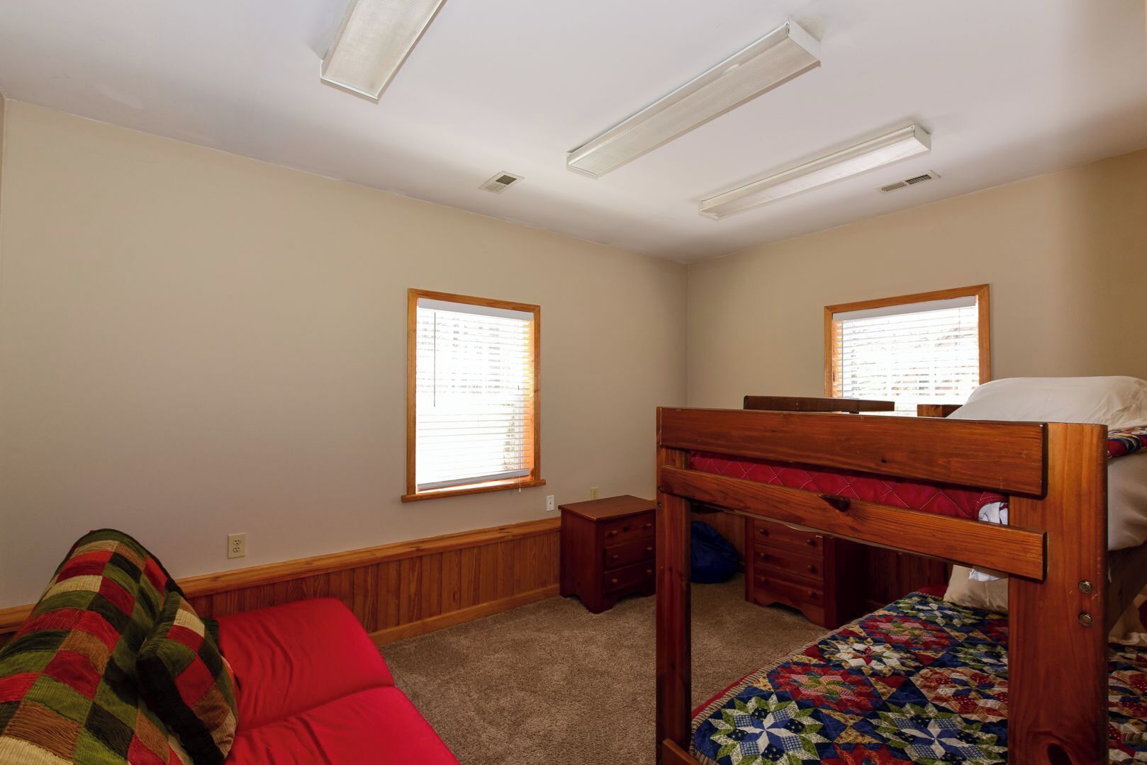 A bedroom with a bunk bed, a single bed, a wooden dresser, a red sofa, and two windows with blinds.