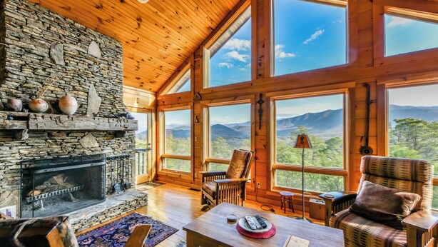 A cozy cabin interior with a stone fireplace, wooden walls, large windows, and a view of mountains.