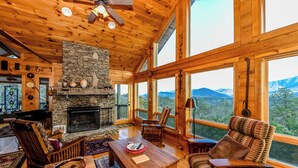 A cozy cabin interior with a stone fireplace, wooden furniture, and large windows offering mountain views.