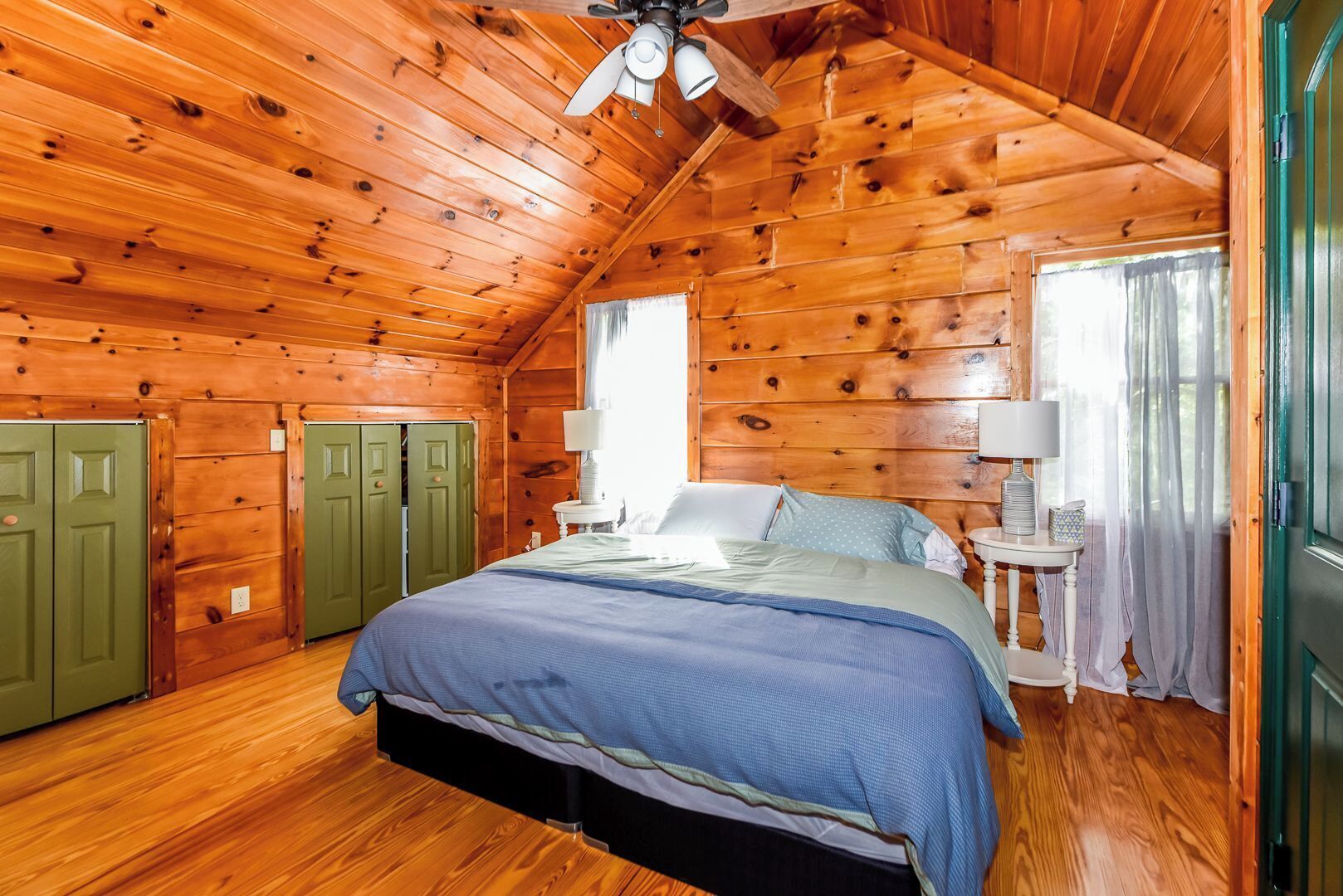 A wooden cabin bedroom with a bed, two green doors, a ceiling fan, and a window with curtains.