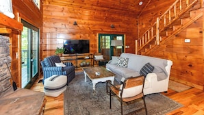 A cozy log cabin interior with a stone wall, wooden floors, a TV, a sofa, and a staircase.