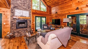 A cozy cabin interior with a stone fireplace, wooden walls, a large window, and a comfortable seating area.