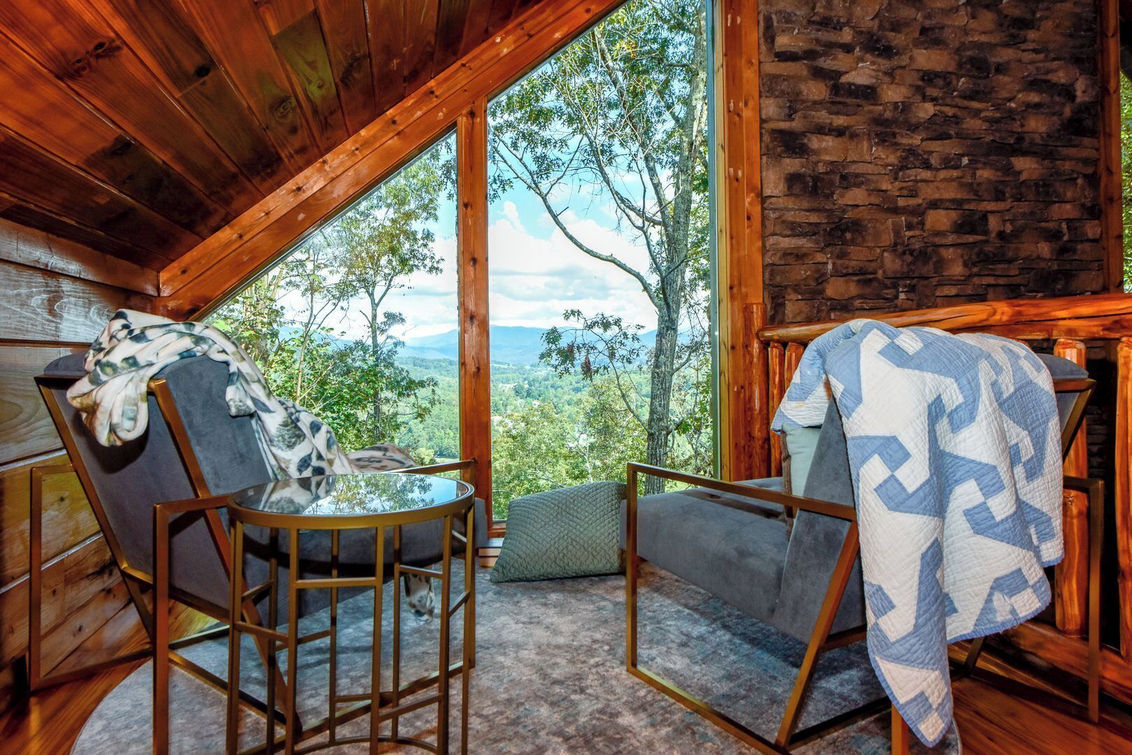 Habitació d'un xalet acollidora amb una finestra gran que ofereix una vista panoràmica d'arbres i muntanyes. Hi ha dues cadires amb mantes, una taula rodona petita i una paret de pedra.