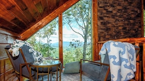 A cozy cabin room with a large window offering a scenic view of trees and mountains. There are two chairs with blankets, a small round table, and a stone wall.