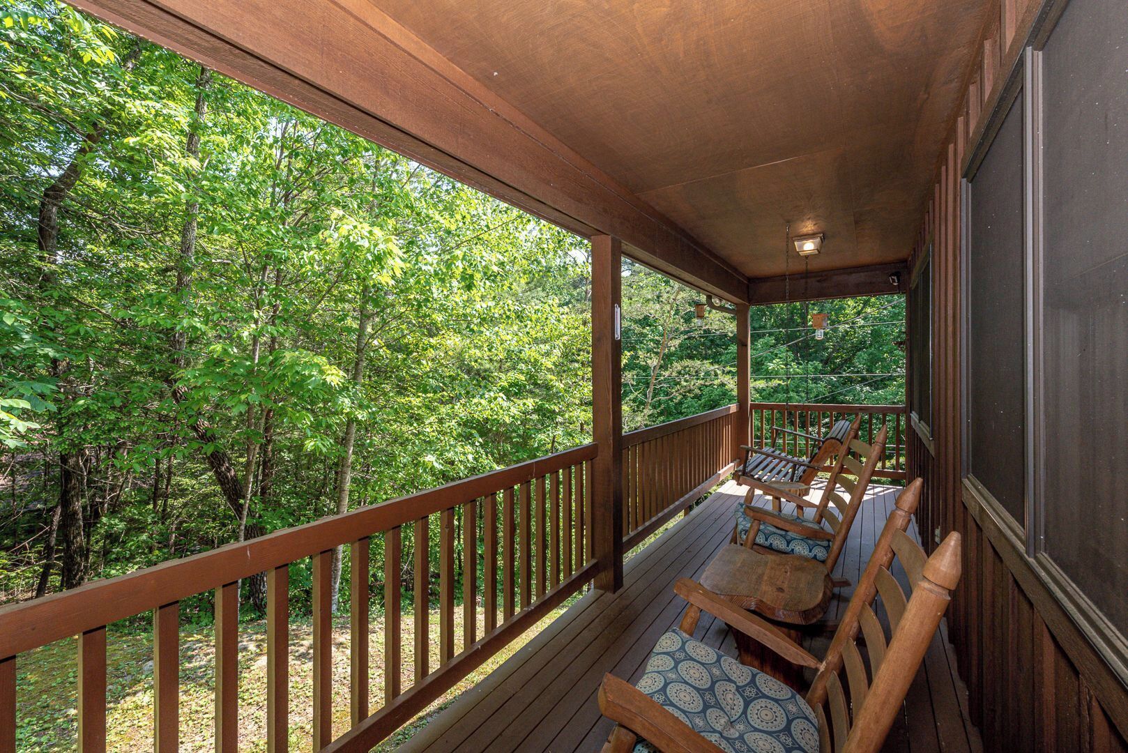 A wooden deck with a railing, two wooden chairs with patterned cushions, and a view of dense green trees.