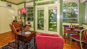 Una habitación acogedora con una mesa de madera, sillas, un sillón mecedora y un escritorio con una lámpara. Amplias ventanas ofrecen vistas al exterior, con zonas verdes.