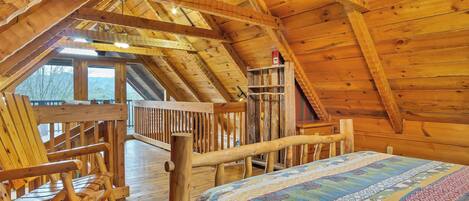 A wooden interior with a high ceiling, a bed with a patterned bedspread, wooden chairs, and a wooden railing.