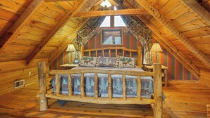 Uma cabana de madeira com quarto no mezanino, com cama e roupa de cama estampada, cabeceira de madeira e janela com cortinas.