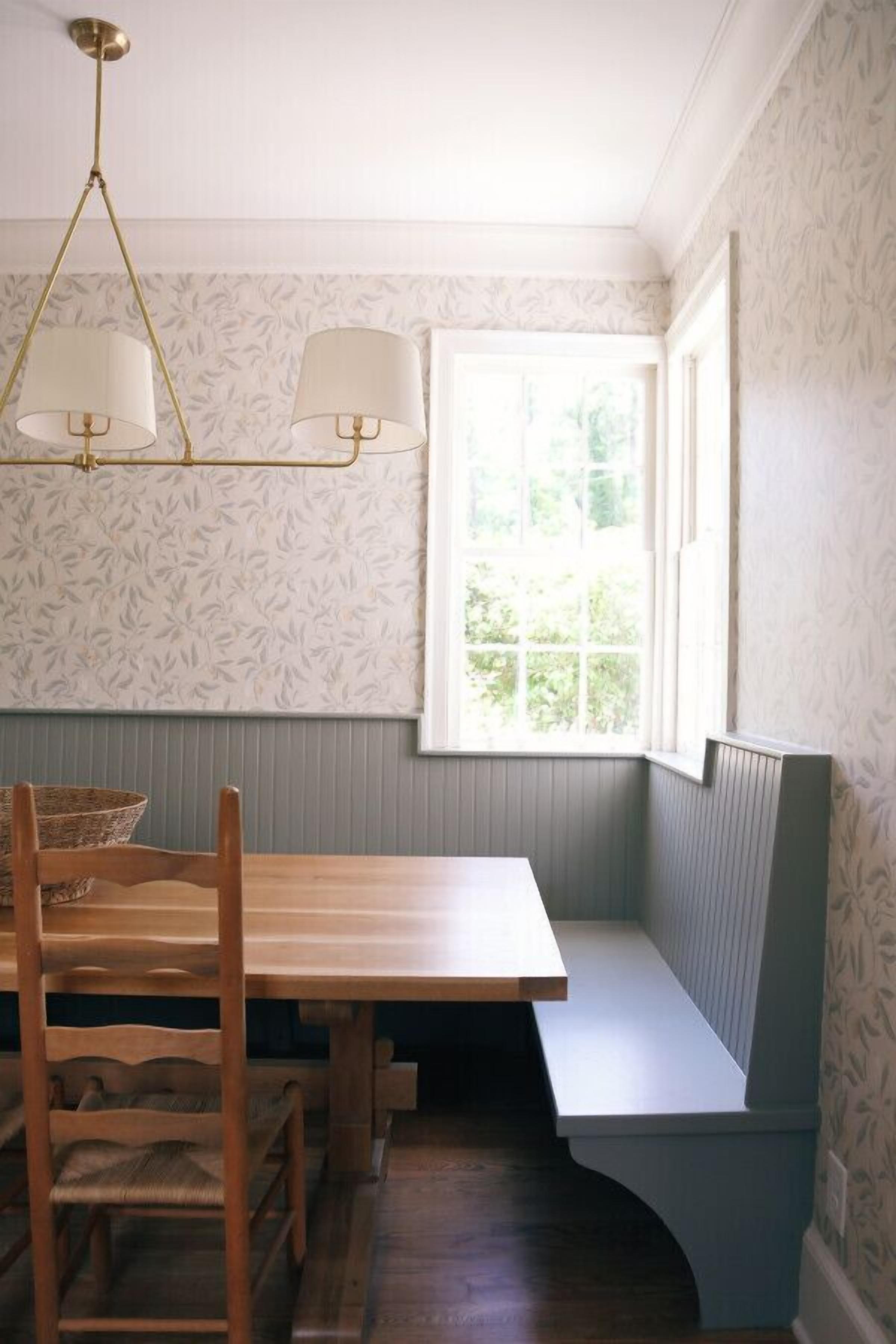 A corner room with a wooden table and bench, a window with a view, and patterned wallpaper.
