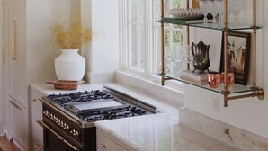 Una cocina con estufa a gas, armarios blancos y repisas de vidrio con objetos decorativos.