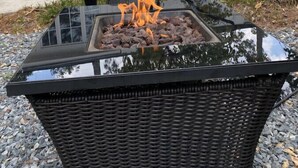 Un hogar con cubierta de vidrio y leña encendida, rodeado de sillas de mimbre, frente a una casa blanca con un árbol.