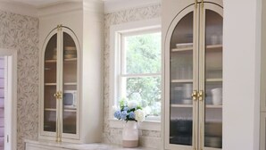 Una cocina con armarios blancos, tiradores dorados y una ventana con un jarrón de flores.