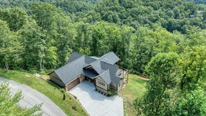 A two-story house with a garage, surrounded by dense forest and mountains in the background.