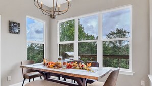 A dining area with a wooden table set for six, surrounded by chairs, large windows with a view of trees, and a chandelier.