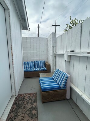 Un petit patio extérieur avec deux canapés en osier dotés de coussins à rayures, un paillasson décoratif en métal et un mur en briques blanches.