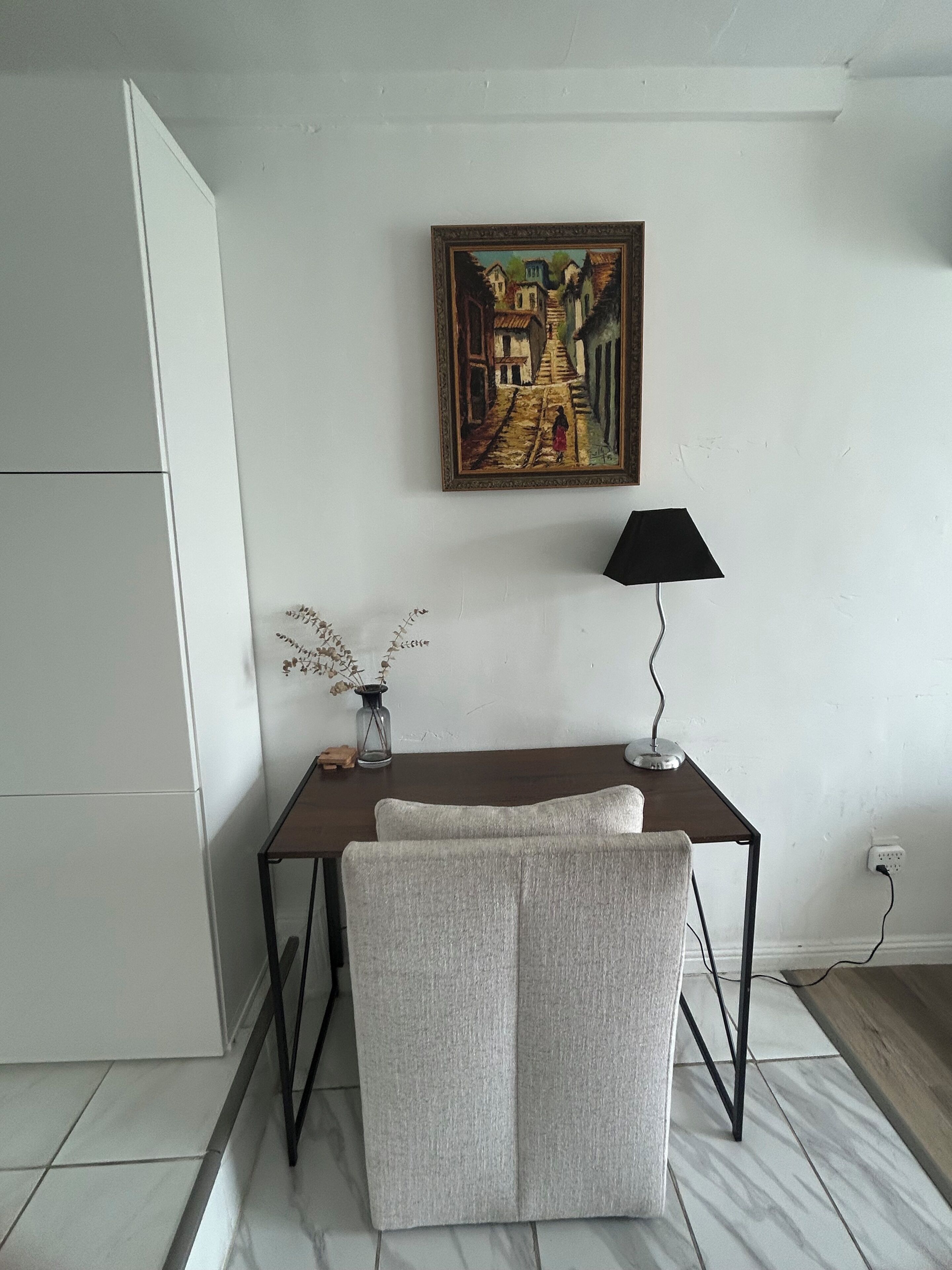 A room with a desk, a chair, a lamp, a painting, and a vase with dried flowers.