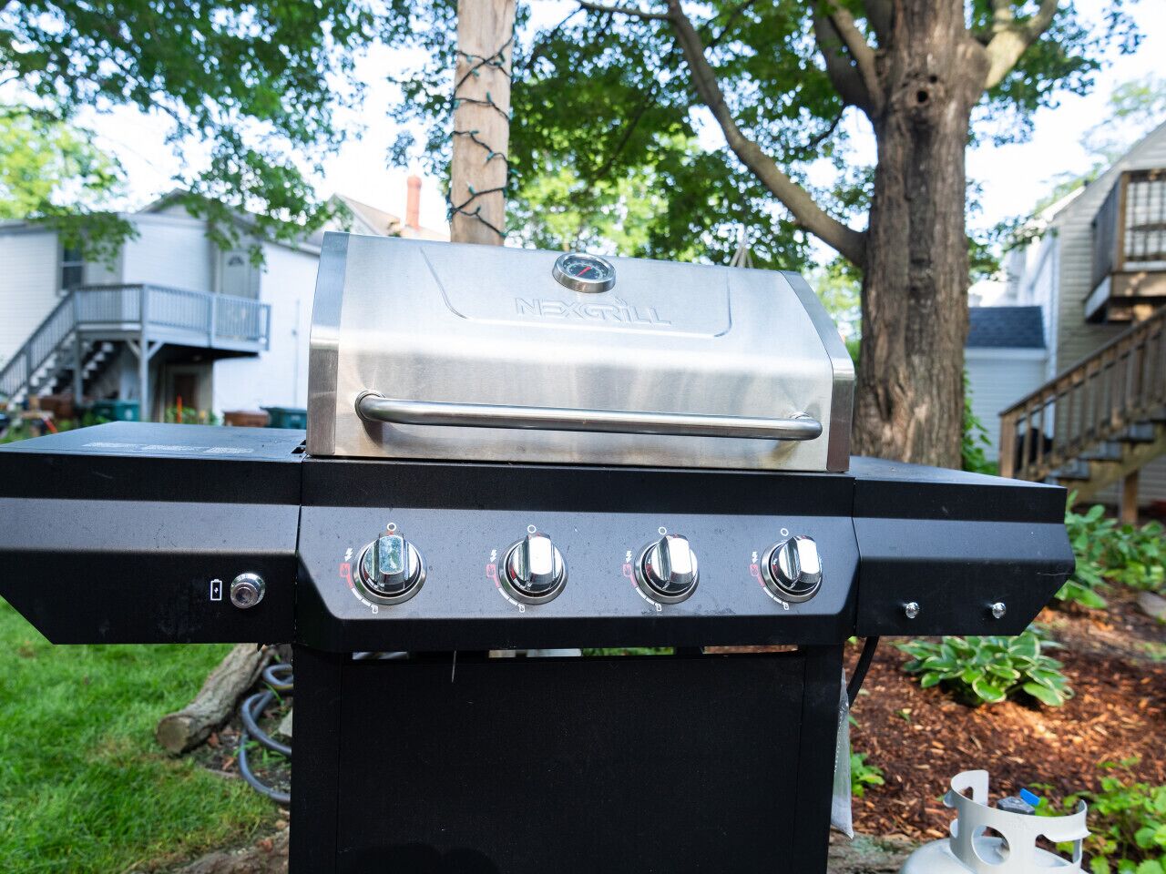 Ein schwarzer Outdoor-Grill mit vier Reglern und einem digitalen Display, vor einer Gartenlandschaft mit Baum und Haus im Hintergrund.