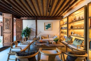 Ein gemütliches Wohnzimmer mit Holzmöbeln, einer Couch und einem Couchtisch. Regale mit Dekorationselementen und ein Topfpflanze sind ebenfalls vorhanden.